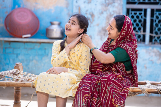 Rural indian mother braids hair of her young adorable daughter, cute girl feel pain as mum getting her ready sitting on traditional bed at village home. Both spending time together.