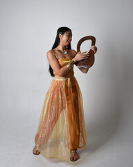 Full length portrait of pretty young asian woman wearing golden Arabian robes like a genie, holding...