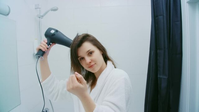 Front view of beautiful woman in white bathrobe hair dryer drying dark hair after shower in modern bathroom. Concept of pretty woman drying hair on white small bathroom.