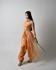 Full length portrait of pretty young asian woman wearing golden Arabian robes like a genie, holding a sword weapon, isolated on studio background.