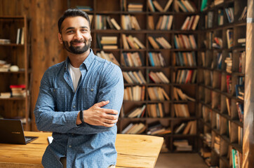 Smiling bearded indian businessman stands near desk and looks at the camera. Young positive male student in library with bookshelves on background. Proud and successful mixed-race small business owner