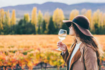 Smiling young woman in stylish hat and coat drinks delicious white wine from wineglass near...