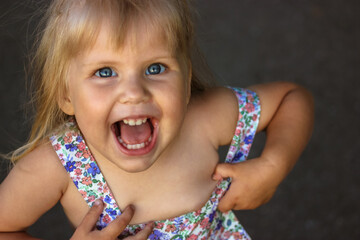 A beautiful cheerful blonde girl of 2-3 years old laughing, looking up. Female child, kid in a flowered sundress playing outdoors at sunny summer day. Fun, joy concept. Happy childhood. Real emotions.