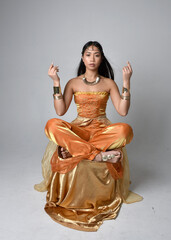 Full length portrait of pretty young asian woman wearing golden Arabian robes like a genie, seated...