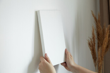Blank canvas in female hands, picture mockup. Woman hanging canvas on white wall