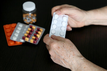 Wrinkled hands of elderly woman with pills on dark wooden table. Different medication in tablets and capsules, taking sedatives, antibiotics or vitamins