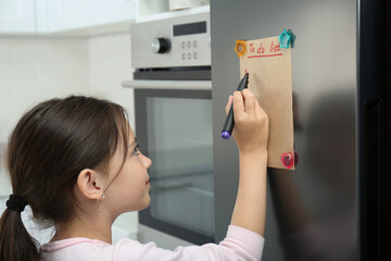 Little girl writing to do list on fridge in kitchen