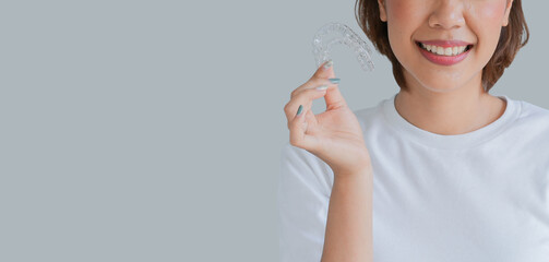 close up young asian woman smiling with hand holding dental aligner retainer (invisible) isolated on gray background of dental clinic for beautiful teeth treatment course concept