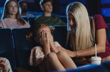 Worried small girl covering eyes in the cinema while watching film, mother comforting her.