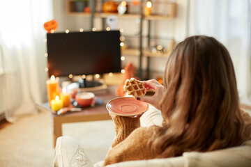 halloween, holidays and leisure concept - young woman watching tv and eating waffle at cozy home