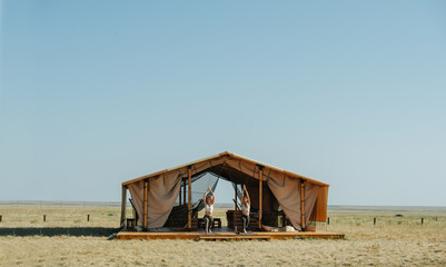 Far shot of two women doing yoga on a doorstep of a huge wooden pole tent