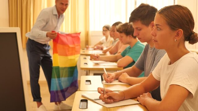 Focused teenage schoolgirl sitting at lesson with classmates, listening young teacher talking about LGBT community and showing rainbow flag. High quality 4k footage