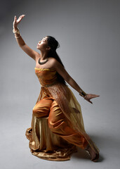 Full length portrait of pretty young asian woman wearing golden Arabian robes like a genie, seated...