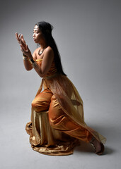 Full length portrait of pretty young asian woman wearing golden Arabian robes like a genie, seated pose, isolated on studio background.