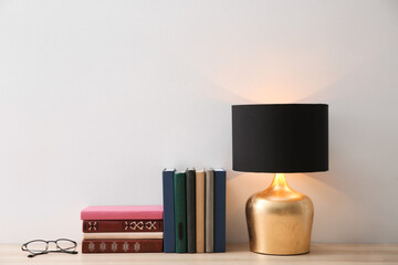 Collection of books, glasses and lamp on wooden table near white wall. Space for text