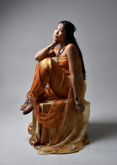 Full length portrait of pretty young asian woman wearing golden Arabian robes like a genie, seated...