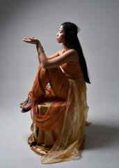 Full length portrait of pretty young asian woman wearing golden Arabian robes like a genie, seated...
