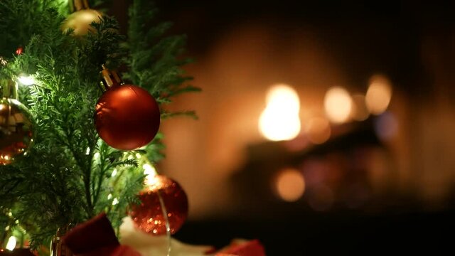 Christmas tree lights by fire in fireplace, New Year or Xmas decoration of pine or fir with red balls. Fireside on winter christmastime holiday. Cozy place by firewood. Seamless looped cinemagraph.