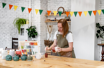 Woman potter in apron making pottery and painting ceramic dishware in workshop. The master potter works in a workshop