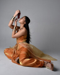 Full length portrait of pretty young asian woman wearing golden Arabian robes like a genie, seated...