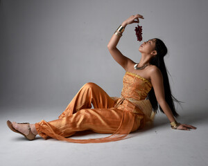 Full length portrait of pretty young asian woman wearing golden Arabian robes like a genie, seated...
