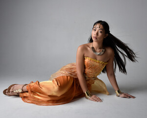 Full length portrait of pretty young asian woman wearing golden Arabian robes like a genie, seated...