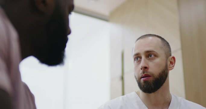 Close-up face a male medical worker is sitting in the dental clinic and explaining something to an African American male patient.