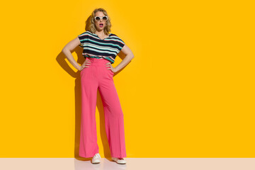 Surprised Young Woman In Sunglasses, Pink Wide Leg Trousers, Sneakers Is Standing With Hands On Hip - 449855615