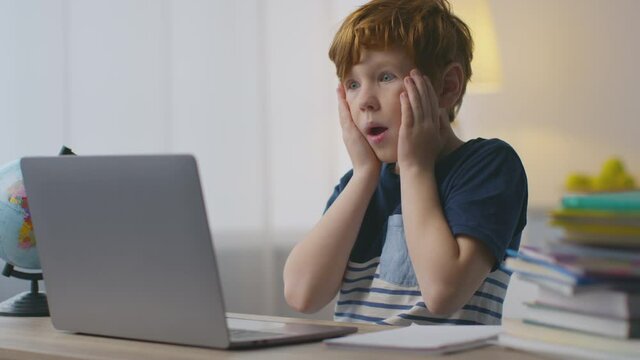Portrait of shocked little redhead boy looking at laptop with amazed expression, touching his cheeks in amazement