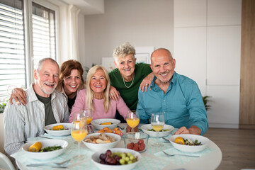 Group of senior friends having party indoors, looking at camera when eating.
