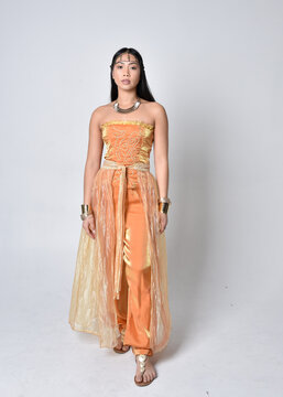 Full length portrait of pretty young asian woman wearing golden Arabian robes like a genie, standing pose  isolated on studio background.