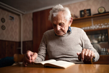 Portrait of elderly man sitting at the table indoors at home, reading book.