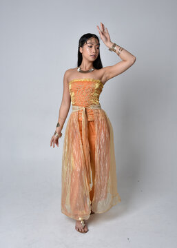 Full length portrait of pretty young asian woman wearing golden Arabian robes like a genie, standing pose  isolated on studio background.