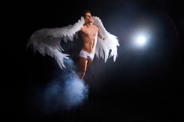 Handsome young athletic man with a bare torso who looks like an angel with white wings. Model dancer posing in a dark studio on black background