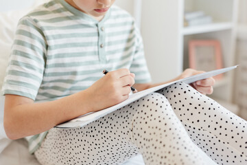 Minimal cropped shot of little girl sitting on bed and drawing in white room, copy space