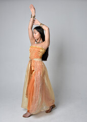 Fototapeta premium Full length portrait of pretty young asian woman wearing golden Arabian robes like a genie, standing pose isolated on studio background.