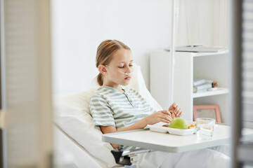 Minimal side view portrait of girl eating healthy lunch while lying on bed in hospital room