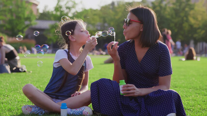 Caucasian preteen daughter and african mother blow soap bubbles on blurry background in park