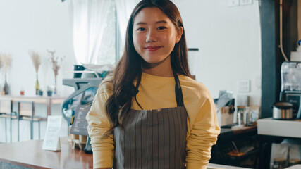 Portrait young Asian woman barista feeling happy smiling at urban cafe. Small business owner Korean girl in apron relax toothy smile looking to camera standing at the counter in coffee shop. - obrazy, fototapety, plakaty