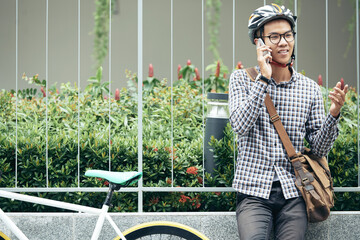 Portrait of positive student in plaid shirt and bicycle helmet talking on phone with friend of...