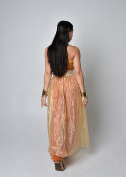 Full length portrait of pretty young asian woman wearing golden Arabian robes like a genie, standing pose  with back to the camera, isolated on studio background.