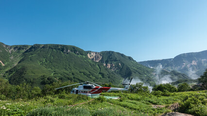 Fototapeta na wymiar The helicopter is standing on the landing pad. There is lush grass and wildflowers in the meadow. There is green vegetation on the mountain slopes. Steam rising from the Valley of Geysers is visible.