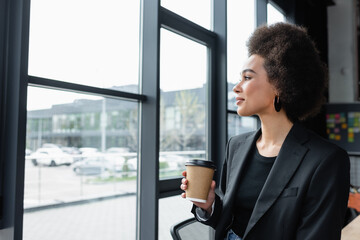 african american businesswoman with takeaway drink looking through window in office