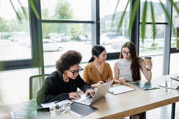 african american businesswoman typing on laptop near interracial colleagues working with documents