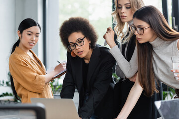 asian businesswoman writing in notebook near multicultural colleagues looking at laptop