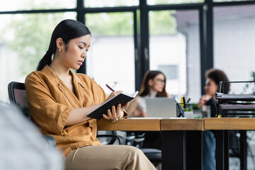 asian businesswoman writing in notebook near multiethnic coworkers on blurred background
