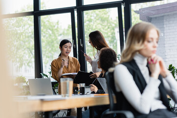 excited asian woman talking to business partner near blurred multiethnic colleagues