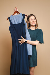 Charming dark blond caucasian woman in green dress smiles widely and holds hanger with dresses dress, trying to decide what to wear.