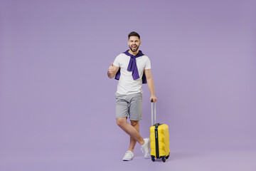 Full size body length side view traveler tourist young brunet man 20s wear white t-shirt purple shirt hold suitcase bag show thumb up like gesture isolated on pastel violet background studio portrait.