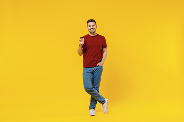 Fototapeta na wymiar Full length smiling happy fun young man 20s wear red t-shirt casual clothes hold takeaway delivery craft paper brown cup coffee to go isolated on plain yellow color wall background studio portrait